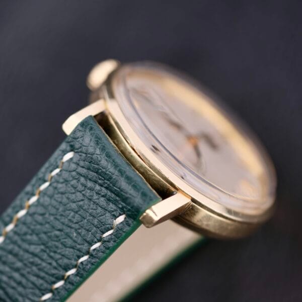 Close-up of an Omega Automatic Day/Date Linen M Dial wristwatch with a green leather strap and white stitching. The gold casing and clear face stand out against a blurred dark background.