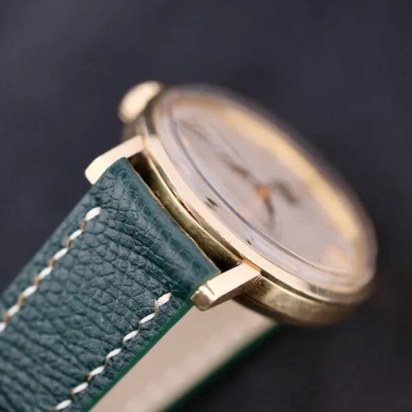 Close-up of an Omega Automatic Day/Date Linen M Dial wristwatch with a green leather strap and white stitching. The gold casing and clear face stand out against a blurred dark background.