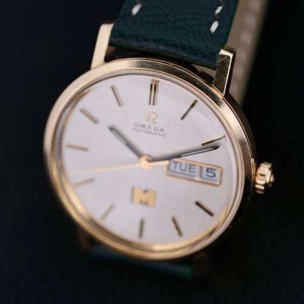A close-up of the Omega Automatic Day/Date Linen M Dial wristwatch reveals a gold case with a black leather strap. The dial, featuring a sophisticated design, includes gold hands and markers with the day TUE and date 5, set against a stark black background.