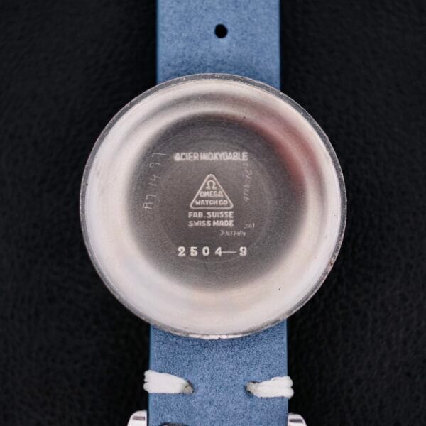 The image shows the back of an Omega Pre Seamaster Cal. 283 watch case with engravings like Acier Inoxydable, Omega Watch Co, Fab. Suisse, and 2504-9. It is paired with a blue strap featuring white stitching against a black background.