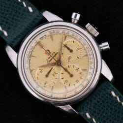 The Omega Seamaster Chronograph KO 2907 is a vintage watch featuring a cream dial, silver case, and three subdials. It has a tachymeter scale on the outer rim and comes with a textured green leather strap and thin metallic hands.