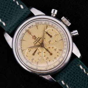 The Omega Seamaster Chronograph KO 2907 is a vintage watch featuring a cream dial, silver case, and three subdials. It has a tachymeter scale on the outer rim and comes with a textured green leather strap and thin metallic hands.
