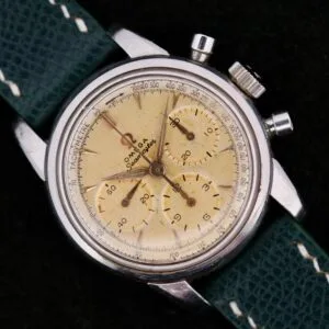 The Omega Seamaster Chronograph KO 2907 is a vintage watch featuring a cream dial, silver case, and three subdials. It has a tachymeter scale on the outer rim and comes with a textured green leather strap and thin metallic hands.