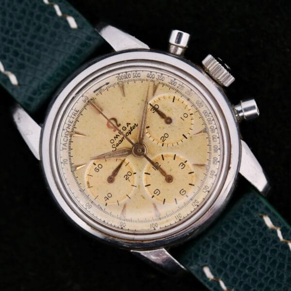 The Omega Seamaster Chronograph KO 2907 is a vintage watch featuring a cream dial, silver case, and three subdials. It has a tachymeter scale on the outer rim and comes with a textured green leather strap and thin metallic hands.