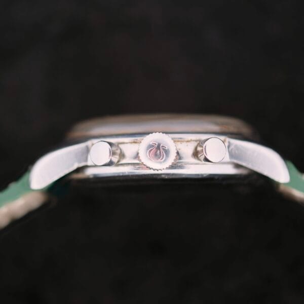 A close-up side view of the Omega Seamaster Chronograph KO 2907 highlights its two pushers and crown on a polished surface, with a partially visible watch band. The intricate details are accentuated by a dark background.