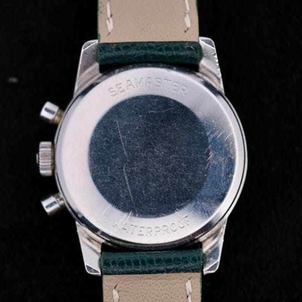 Back view of a vintage Omega Seamaster Chronograph KO 2907 wristwatch with cream and dark green leather strap, words Seamaster and Waterproof engraved on the metal casing. Scratches on the surface hint at its storied past.