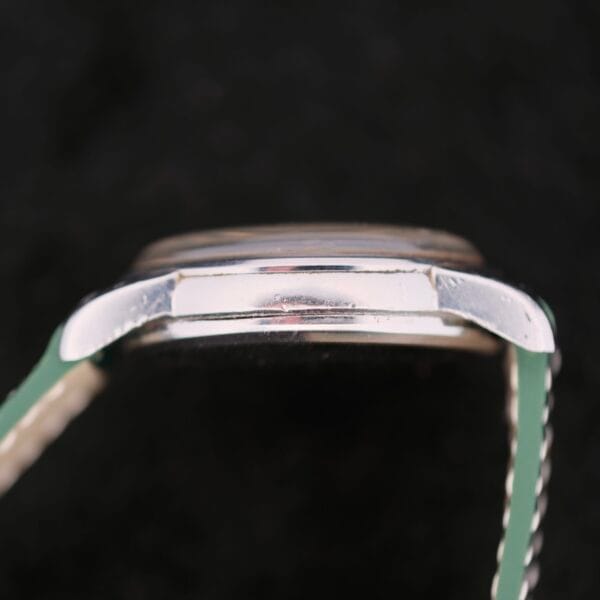 Close-up side view of a silver watch case against a black background, highlighting the thin, polished edge reminiscent of the Omega Seamaster Chronograph KO 2907 collection. A slightly blurred green strap with white stitching is visible.