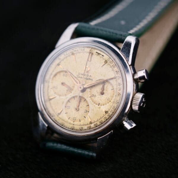 Close-up of a vintage Omega Seamaster Chronograph KO 2907 watch with a worn beige dial, silver case, dark green leather strap, and three sub-dials on a dark surface.