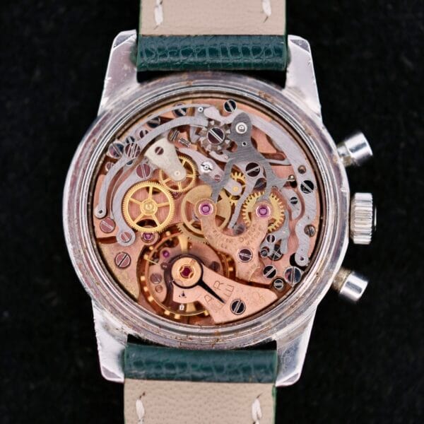 Close-up of an Omega Seamaster Chronograph KO 2907 with the back case removed, displaying intricate gears. It features a silver case and a beige leather strap with green edges, set against a black background.