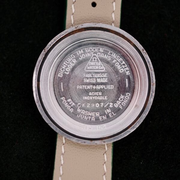 Close-up of the back of an Omega Seamaster Chronograph KO 2907 watch case, showcasing engraved text like OMEGA WATCH CO and FAB. SUISSE SWISS MADE. The watch case rests on a light-colored strap against a dark background.