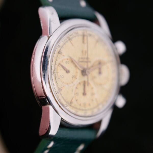 A close-up of the Omega Seamaster Chronograph KO 2907 wristwatch showcases its round, weathered cream dial with three sub-dials, stainless steel case, and a dark green leather strap with white stitching.