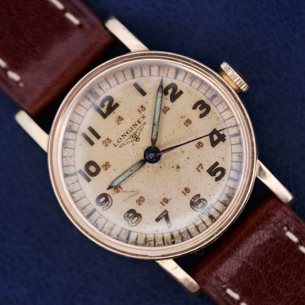 Longines Military 24 Hour Dial 12L wristwatch with a brown leather strap and an aged cream dial, showcased on a blue background.