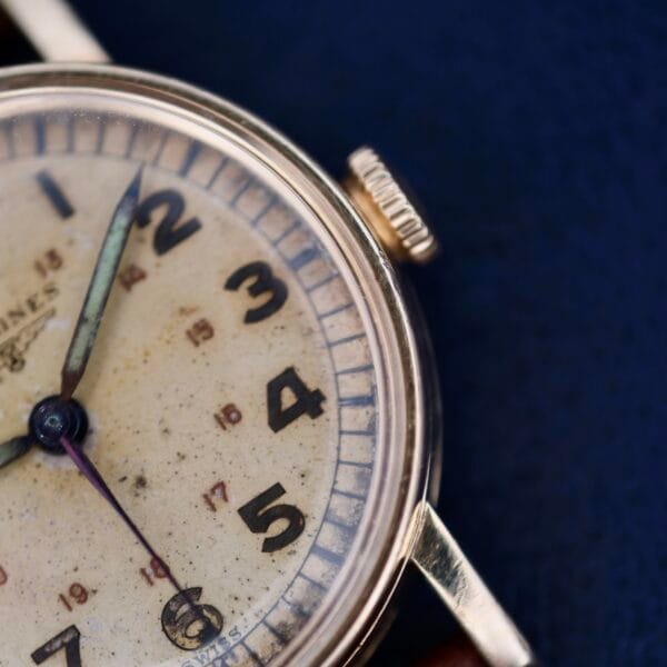 Close-up of a vintage Longines Military 24 Hour Dial 12L wristwatch on a blue background, featuring a worn, discolored dial with large black numerals and dark hands, with its crown prominently visible on the side.