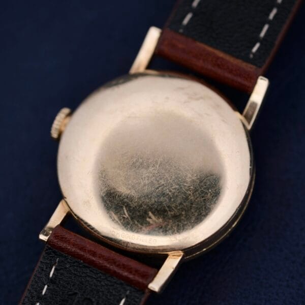 The Longines Military 24 Hour Dial 12L wristwatch, featuring a gold-filled back and a brown leather strap, sits elegantly against a dark blue background. The worn metal surface and scratches add to its charm.