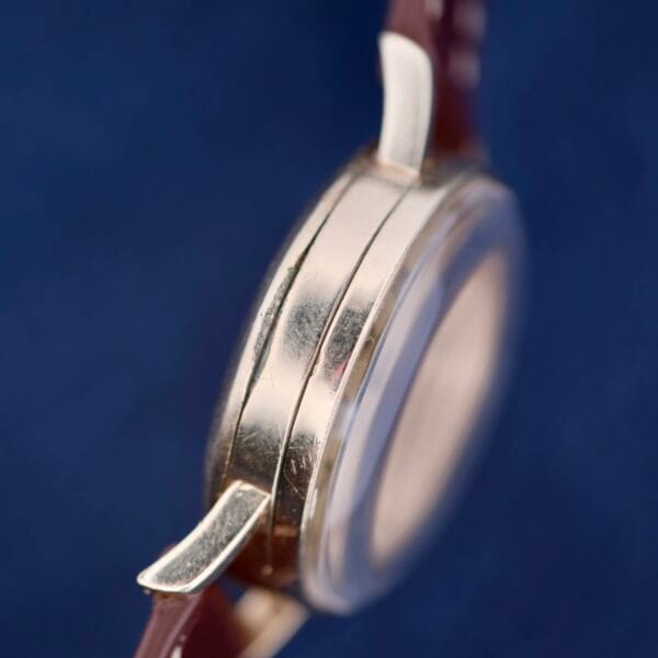 Close-up side view of the Longines Military 24 Hour Dial 12L with a transparent face and two metal lugs, featuring a silver case and brown leather strap. The elegant timepiece stands out against a blurred blue background.