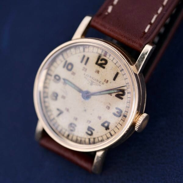 A vintage Longines Military 24 Hour Dial 12L watch features a worn beige dial with black numerals and hands. Its gold-toned case complements the brown leather strap, all set against a softly blurred blue surface.