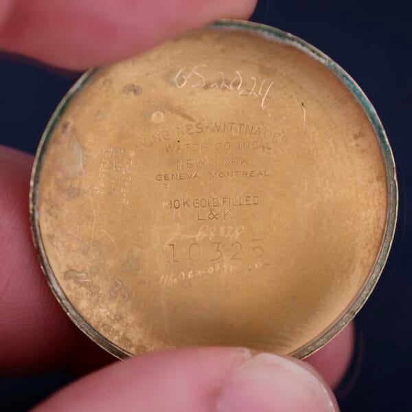 Close-up of a Longines Military 24 Hour Dial 12L watch case back with engraving: LONGINES-WITTNAUER WATCH CO INC. NEW YORK, GENEVA MONTREAL and 10K GOLD FILLED L&K 10325, partially held by two fingers, showcasing its refinement.