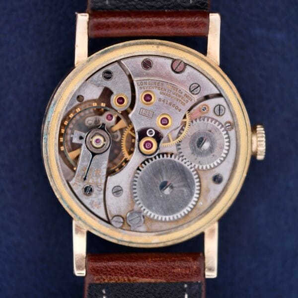 Close-up of the vintage Longines Military 24 Hour Dial 12L watch mechanism, highlighting intricate gears and components. It features a metal frame with visible screws and a brown leather strap, elegantly set against a dark blue background.