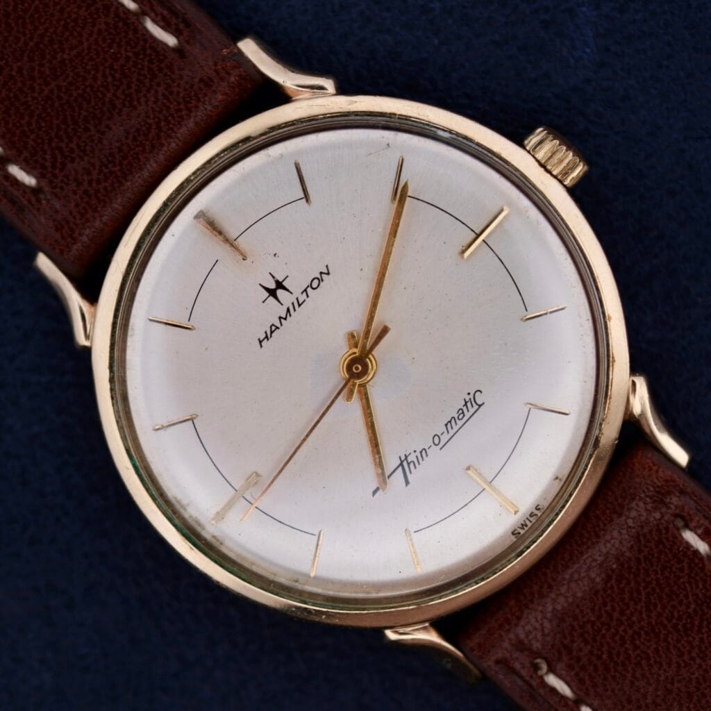 The Hamilton Thin-O-Matic Sunburst wristwatch features a sunburst dial and brown leather strap, shown against a dark blue background.