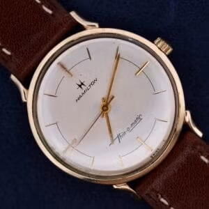 The Hamilton Thin-O-Matic Sunburst wristwatch features a sunburst dial and brown leather strap, shown against a dark blue background.