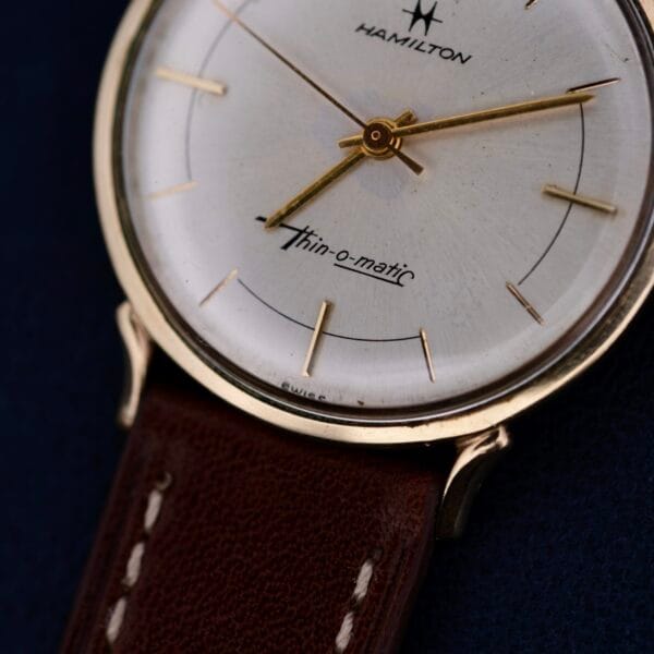 A close-up of the Hamilton Thin-O-Matic Sunburst wristwatch reveals its vintage minimalist design with a gold-tone case, brown leather strap, and gold hands and hour markers against a silver sunburst dial, featuring the iconic Hamilton logo.