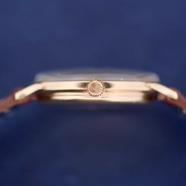 Close-up of a Hamilton Thin-O-Matic Sunburst wristwatch, showcasing the gold side crown against a blurred blue backdrop. The watch features a sleek profile with a shiny, sunburst finish.