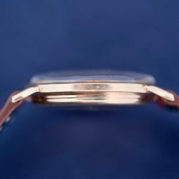 A side view of the Hamilton Thin-O-Matic Sunburst wristwatch against a dark blue background. It features a brown leather strap, and the focus is on its polished gold case with a smooth surface and subtle sunburst effect.