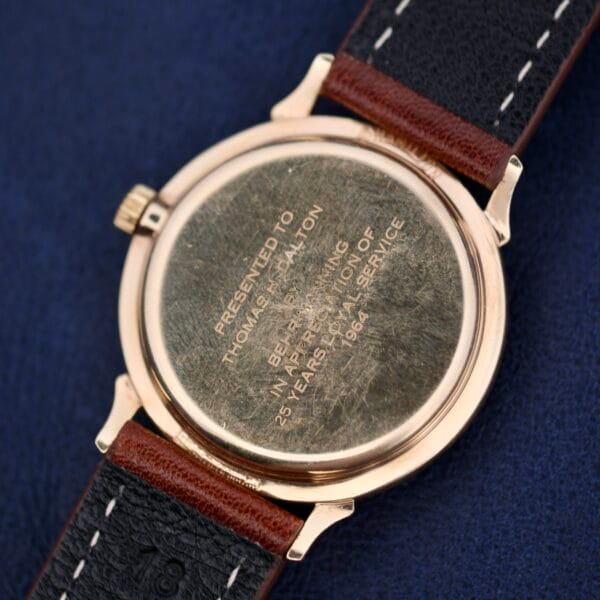 The image shows the back of a Hamilton Thin-O-Matic Sunburst wristwatch, engraved with Presented to Thomas J. Milton in appreciation of 25 years of service, 1964. This timeless piece has a gold finish and includes a black and brown leather strap.