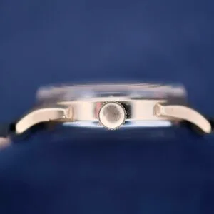 Close-up of a Benrus Day/Date Pointer watch from the side, featuring a silver metal case and crown against a blurred dark blue background. The straps are partially visible on each side.