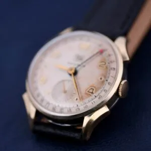 The Benrus Day/Date Pointer analog wristwatch features a white dial with gold numerals and two subdials on a dark strap. The gold-toned case enhances its elegance against the dark blue surface.