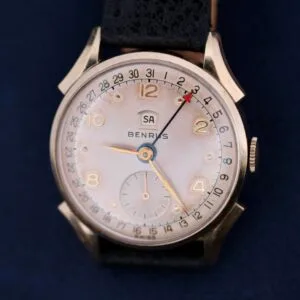 A vintage Benrus Day/Date Pointer wristwatch is showcased, featuring a white face with gold numbers and hands. It includes a small seconds subdial and a date indicator, highlighted by a red arrow at 3, all elegantly set on a dark band.