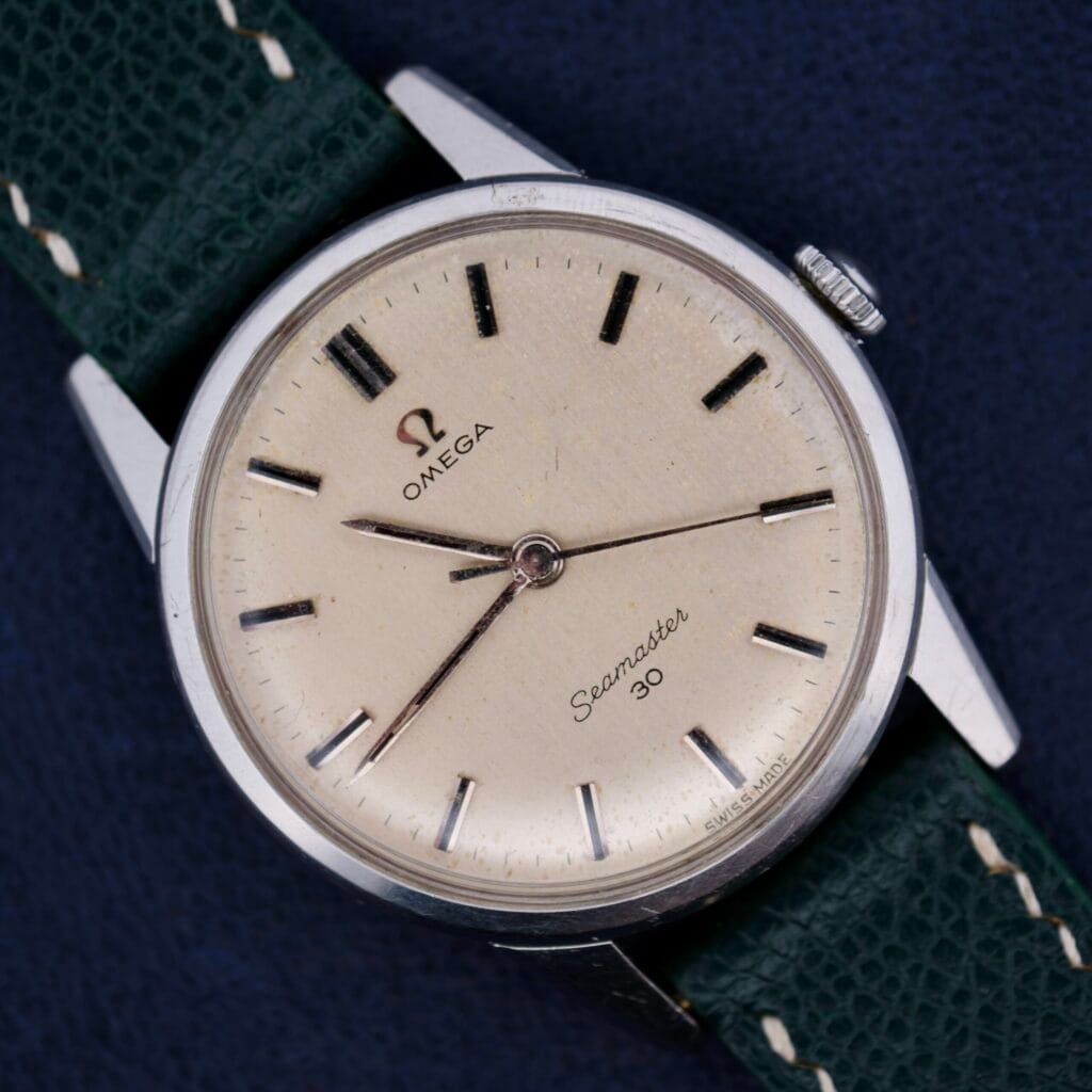 An Omega Seamaster 30 Ref 135.006 with a cream dial and green leather strap rests elegantly on a dark surface.