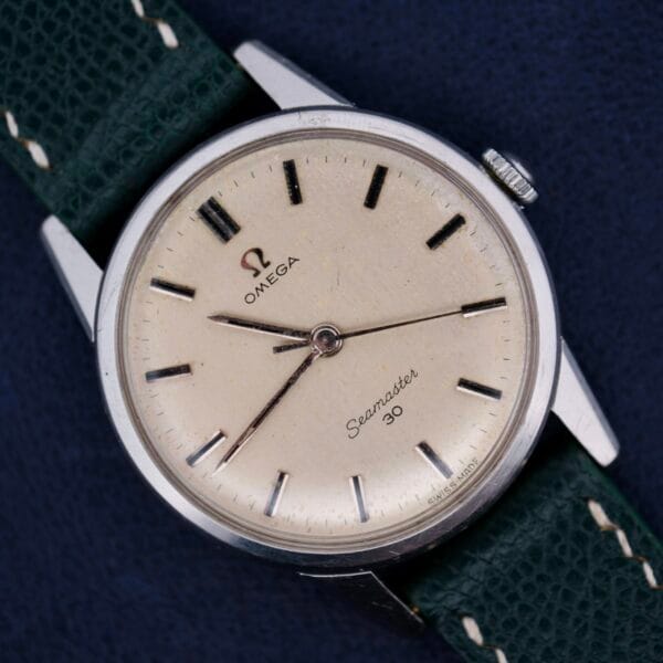 The classic Omega Seamaster 30 Caliber 286 wristwatch features a cream dial with black hour markers, encased in sleek silver. It is elegantly set on a green leather strap with white stitching, all strikingly displayed against a dark blue background.