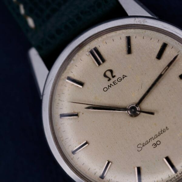 A close-up of the Omega Seamaster 30 Caliber 286 watch reveals a cream dial with sleek black markers and hands. Its silver case and textured dark green strap elegantly contrast against a dark backdrop, showcasing the iconic classic charm of this timepiece.