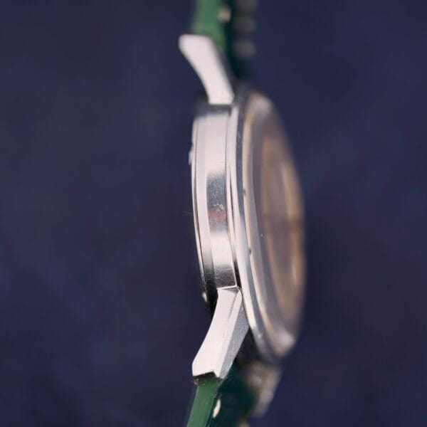 Side view of an Omega Seamaster 30 Caliber 286, highlighting its silver round case and green strap against a dark blue backdrop. The focus is on the metallic case and lugs, while the watch face is artfully blurred.