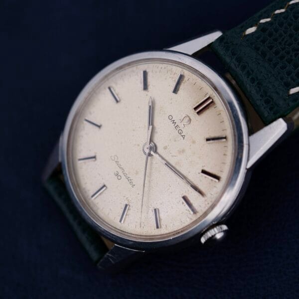 A close-up of the Omega Seamaster 30 Caliber 286 highlights its cream dial and silver hour markers, perfectly complemented by a dark green leather strap, set against a rich, dark background.
