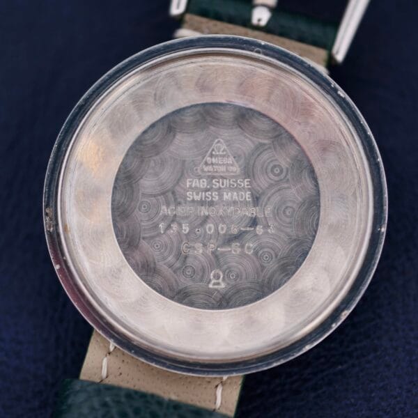 Close-up of the Omega Seamaster 30 Caliber 286 interior case back, showing engravings like FAB. SUISSE, SWISS MADE, and ACIER INOXYDABLE, plus model numbers. The watch has a dark green strap on a dark blue fabric backdrop.