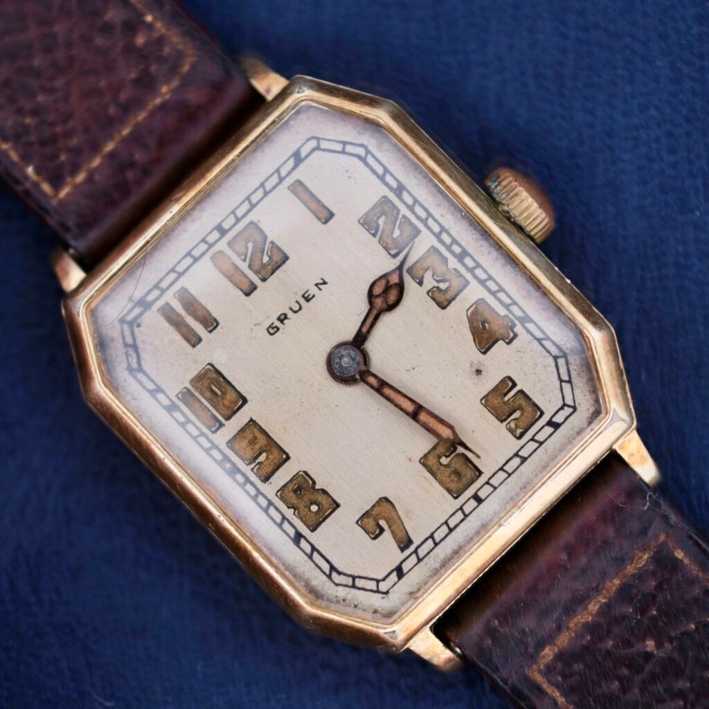 Gruen Guild Art Deco Tank 14k wristwatch featuring a square gold-tone case, brown leather strap, and bold numerals on the dial.
