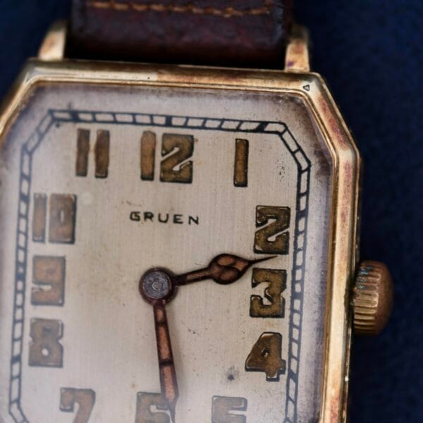 A close-up of the Gruen Guild Art Deco Tank 14k wristwatch showcases its rectangular gold-toned case and leather strap. The off-white face features bold Arabic numerals with ornate dark hands. This vintage piece displays wear and age, enhancing its timeless charm.
.