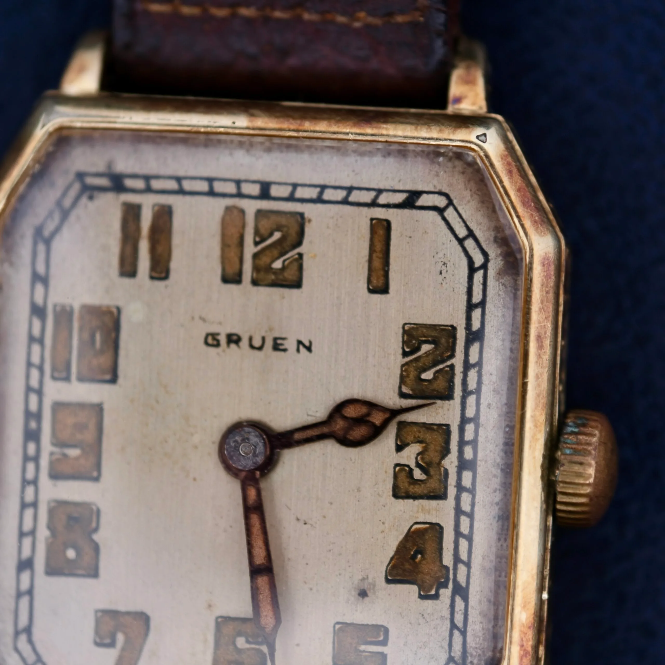 A close-up of the Gruen Guild Art Deco Tank 14k wristwatch showcases its rectangular gold-toned case and leather strap. The off-white face features bold Arabic numerals with ornate dark hands. This vintage piece displays wear and age, enhancing its timeless charm.
.