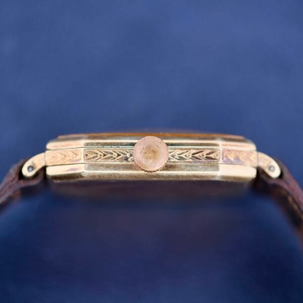Close-up side view of a Gruen Guild Art Deco Tank 14k watch featuring an ornate, engraved gold casing, single button, and brown leather strap set against a smooth dark blue background.