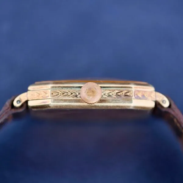 Close-up side view of a Gruen Guild Art Deco Tank 14k watch featuring an ornate, engraved gold casing, single button, and brown leather strap set against a smooth dark blue background.