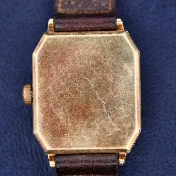 A close-up of a Gruen Guild Art Deco Tank 14k rectangular gold watch reveals scratches and wear on its metal surface, complemented by a dark brown leather strap. The backdrop is textured in dark blue.
