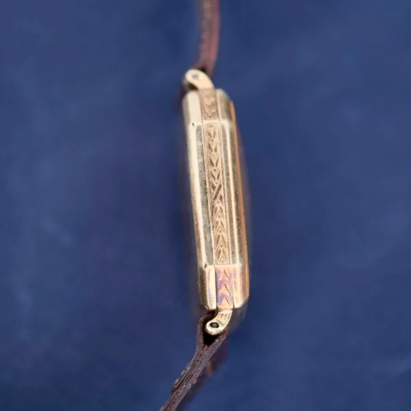 The Gruen Guild Art Deco Tank 14k wristwatch features intricate side engravings and a leather strap, set against a deep blue background that enhances its warm gold tones.