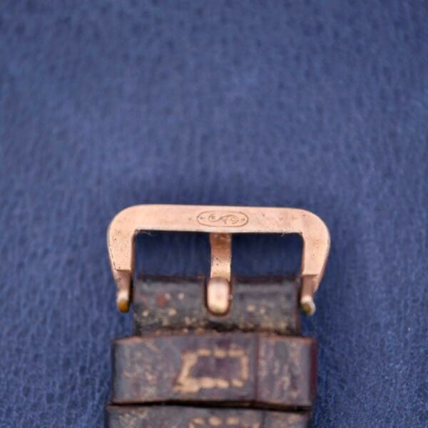 Close-up of a worn leather watch strap with a brass buckle, featuring the engraved Gruen Guild Art Deco Tank 14k logo. The buckle contrasts with a textured blue background.