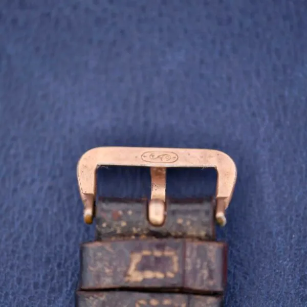 Close-up of a worn leather watch strap with a brass buckle, featuring the engraved Gruen Guild Art Deco Tank 14k logo. The buckle contrasts with a textured blue background.