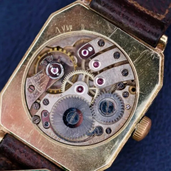 Close-up of a Gruen Guild Art Deco Tank 14k vintage watch movement displaying intricate gears and cogs. The watch features a bronze-toned casing with a leather strap, set against a dark blue background.