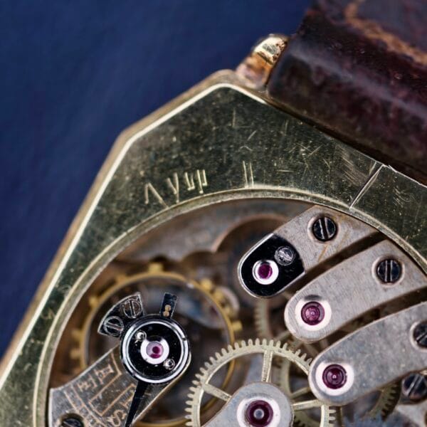 Close-up of a Gruen Guild Art Deco Tank 14k watch showcasing its vintage mechanical design with exposed gears. The aged metal finish complements purple jewels in the mechanism, adding color. The watch strap is visible at the top.