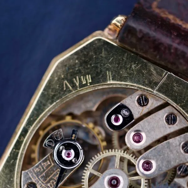 Close-up of a Gruen Guild Art Deco Tank 14k watch showcasing its vintage mechanical design with exposed gears. The aged metal finish complements purple jewels in the mechanism, adding color. The watch strap is visible at the top.