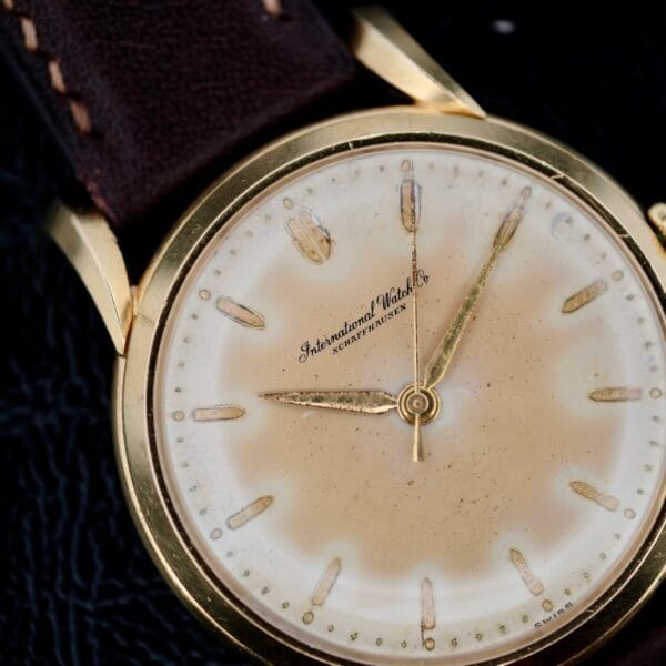 The Iwc Caltrava C. 89 18k Jumbo Case wristwatch exudes timeless elegance with its gold case, brown leather strap, and slightly worn face featuring gold hour markers and hands. The International Watch Co. logo adorns the dial against a dark, textured background that enhances its classic allure.